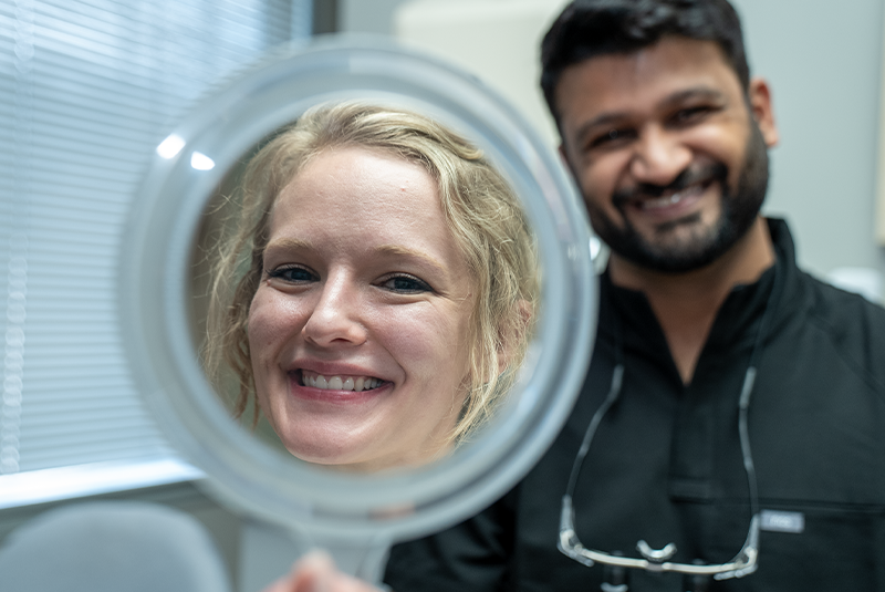 Dr. Bhalerao smiling in the background as patient Ashley smiles into a mirror